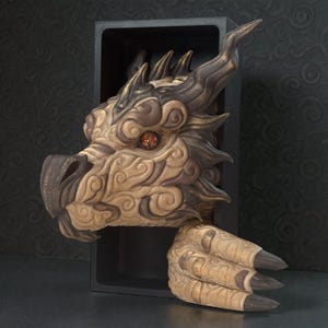May include: A detailed sculpture of a dragon's head and claw emerging from a black box. The dragon features intricate carvings, a tan and brown color scheme, and a fiery red eye. The claw has sharp black talons.