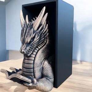 May include: A gold-colored dragon bookend holding a book. The dragon is positioned inside a black box, which is also a bookend.