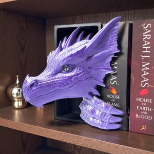 May include: A purple dragon head bookend with textured scales and a detailed eye. The dragon's head is positioned to appear as if it is emerging from a black bookend. The bookend is on a wooden shelf with books.