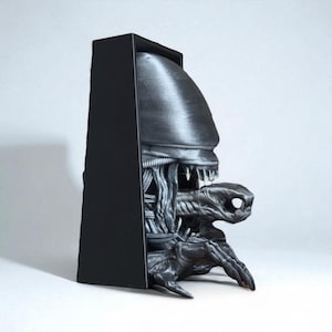 May include: A black and gray sculpture of an Alien Xenomorph emerging from a black rectangular frame. The sculpture features detailed textures and sharp teeth, showcasing the creature's menacing design. The overall theme is science fiction.