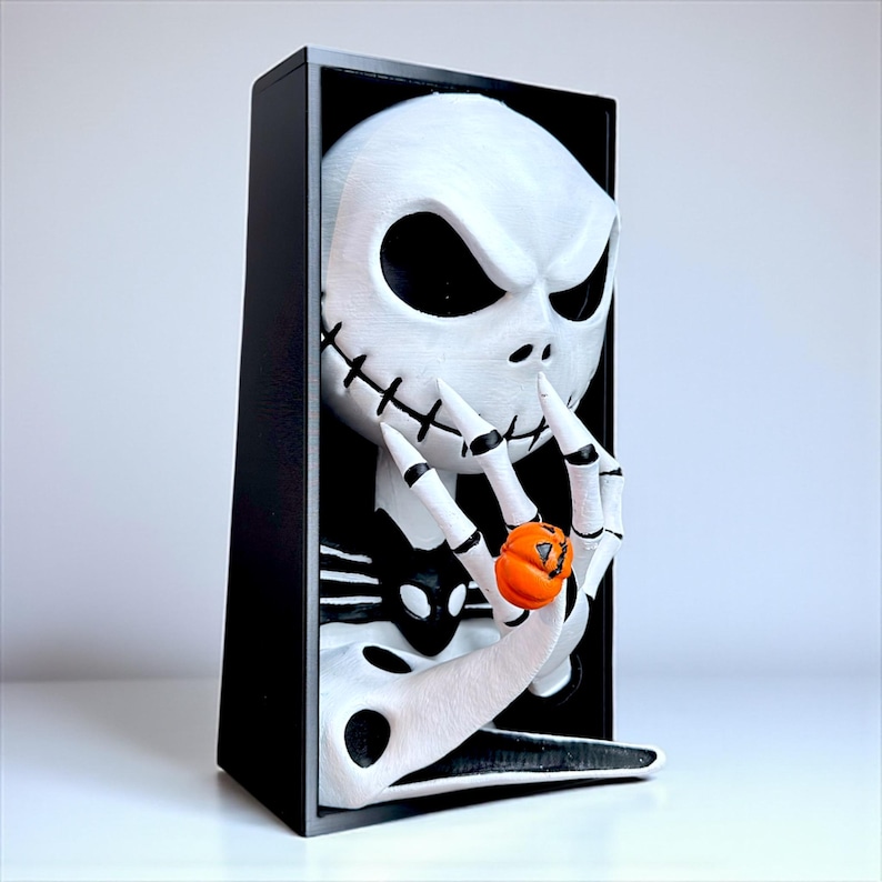 May include: A 3D-printed sculpture of Jack Skellington from The Nightmare Before Christmas. The white skull face with black eyes and stitched mouth is holding an orange pumpkin. The sculpture is set in a black rectangular frame.