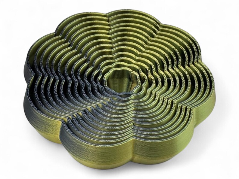 May include: An abstract sculpture in olive green and dark grey, resembling a flower. The piece is composed of concentric, wavy layers, creating a textured surface. The design is modern and artistic, with a unique, layered aesthetic.
