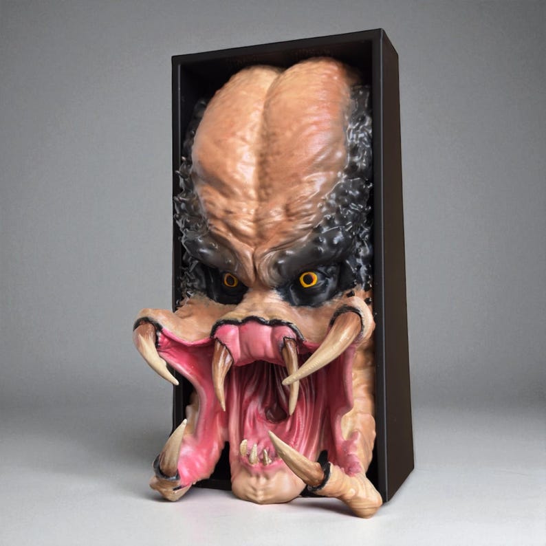 May include: A detailed sculpture of a Predator alien head, featuring a tan face with black and yellow eyes, large teeth, and a wide-open mouth. The sculpture is set within a black rectangular frame.