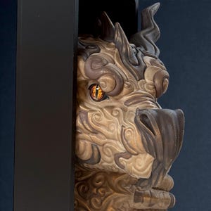May include: A detailed sculpture of a dragon's head, featuring intricate textures and swirling patterns in shades of tan, brown, and gold. The dragon has a piercing orange eye and is partially framed by a black border, creating a dramatic effect.