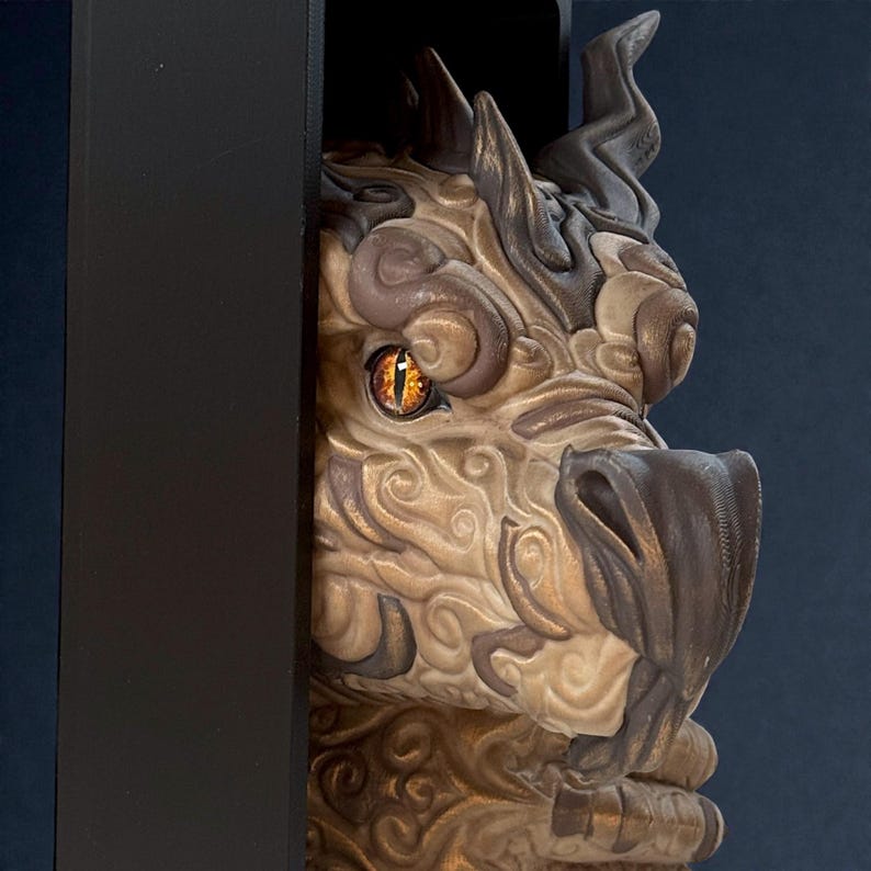 May include: A detailed sculpture of a dragon's head, featuring intricate textures and swirling patterns in shades of tan, brown, and gold. The dragon has a piercing orange eye and is partially framed by a black border, creating a dramatic effect.