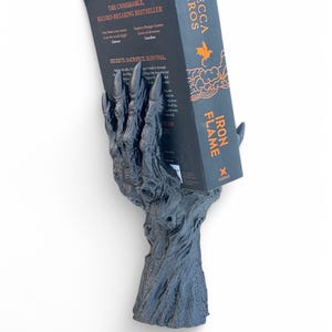 May include: A dark gray, claw-shaped book holder supporting a book titled "Iron Flame" by Rebecca Yarros. The book holder has a textured, tree-like base and is set against a white background. The book's cover is black with orange text.