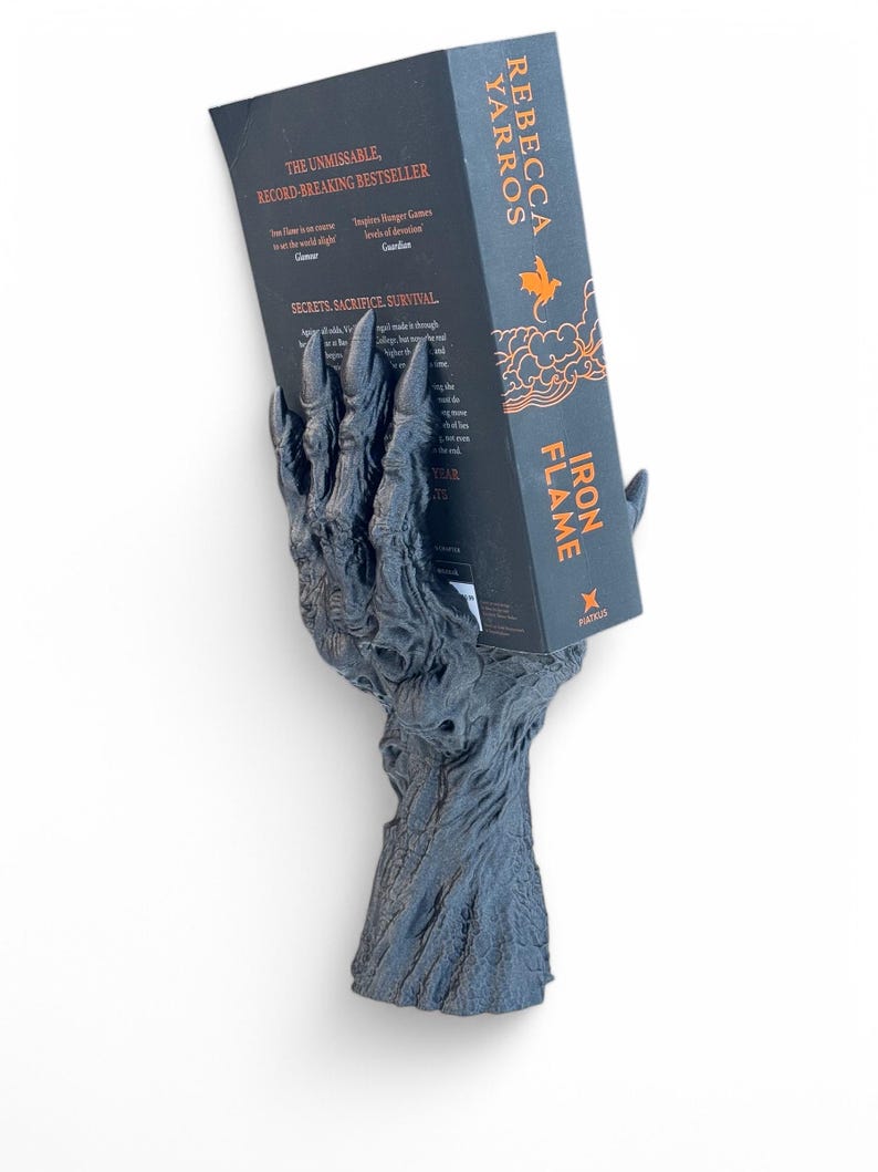 May include: A dark gray, claw-shaped book holder supporting a book titled "Iron Flame" by Rebecca Yarros. The book holder has a textured, tree-like base and is set against a white background. The book's cover is black with orange text.