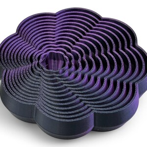 May include: A layered, flower-shaped object in dark purple and black. The design features concentric, wavy layers, creating a three-dimensional effect. The object is likely a decorative item or a mold, possibly for baking.