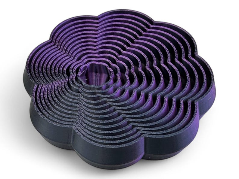 May include: A layered, flower-shaped object in dark purple and black. The design features concentric, wavy layers, creating a three-dimensional effect. The object is likely a decorative item or a mold, possibly for baking.