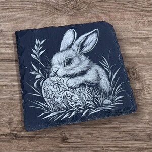 Engraved slate coaster featuring a detailed illustration of a bunny resting on a decorative Easter egg, surrounded by grass and leaves. The coaster has a natural, rough-edged finish and is set on a wooden background