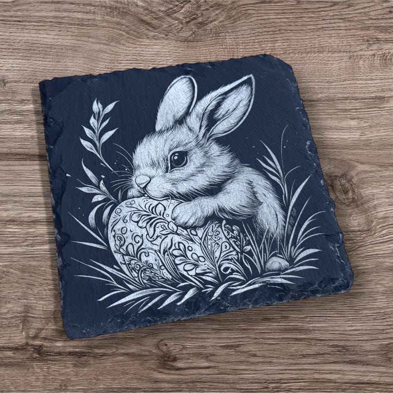 Engraved slate coaster featuring a detailed illustration of a bunny resting on a decorative Easter egg, surrounded by grass and leaves. The coaster has a natural, rough-edged finish and is set on a wooden background