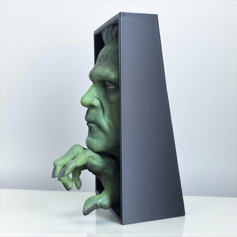 May include: A green Frankenstein's monster bust emerges from a black rectangular frame. The monster's face and hand are detailed with realistic features. The sculpture is set against a white background, creating a striking contrast.