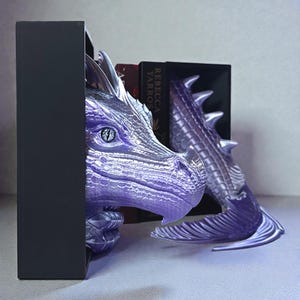 May include: A purple dragon bookend set. The dragon head and tail are detailed with scales and a metallic finish. The bookends are designed to hold books, with a black rectangular base.