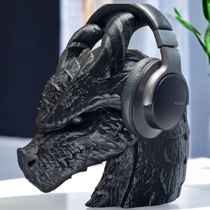 K&ouml;nnte beinhalten: Eine schwarze Drachenkopfskulptur mit einem schwarzen Kopfh&ouml;rer darauf. Der Kopfh&ouml;rer hat einen silbernen Akzent und das Wort "Audio-Technica" ist auf der Seite aufgedruckt.