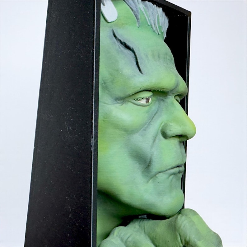 May include: A green Frankenstein monster bust with a black frame. The bust is painted in shades of green and gray, with detailed facial features. The monster's hand is visible, resting on the frame.