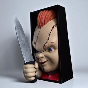 Puede incluir: Escultura de mu&ntilde;eco Chucky con un cuchillo. El mu&ntilde;eco tiene pelo rojo, ojos azules y rasgos faciales cosidos. Est&aacute; colocado en una caja negra, con la mano del mu&ntilde;eco sosteniendo un cuchillo plateado.