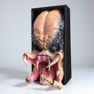May include: A detailed sculpture of a Predator alien head, emerging from a black rectangular frame. The head features a tan forehead, black and textured hair, yellow eyes, and large, sharp teeth. The mouth is open, revealing a pink interior.