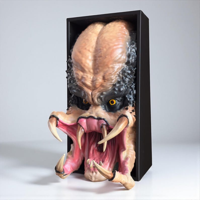 May include: A detailed sculpture of a Predator alien head, emerging from a black rectangular frame. The head features a tan forehead, black and textured hair, yellow eyes, and large, sharp teeth. The mouth is open, revealing a pink interior.