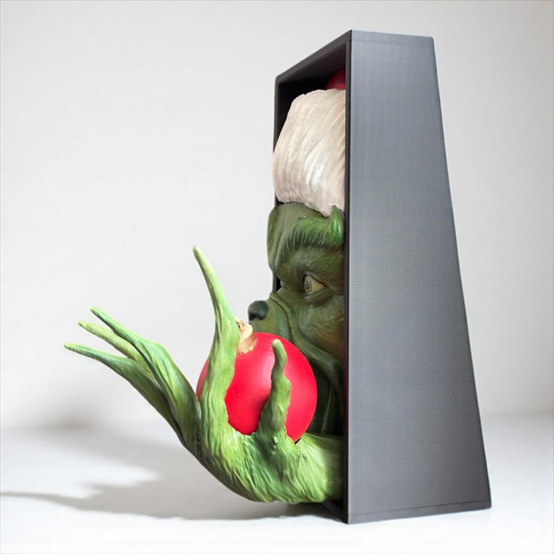 May include: A green Grinch figure holding a red ornament, emerging from a gray rectangular frame. The Grinch's face and hand are visible, with a white Santa hat. The ornament is held in the Grinch's green hand.