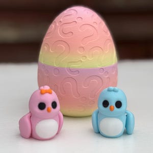 May include: Two small plastic toy birds, one pink and one blue, with white bellies and orange beaks. The birds are sitting in front of a larger pink and yellow egg-shaped toy with question marks on it.