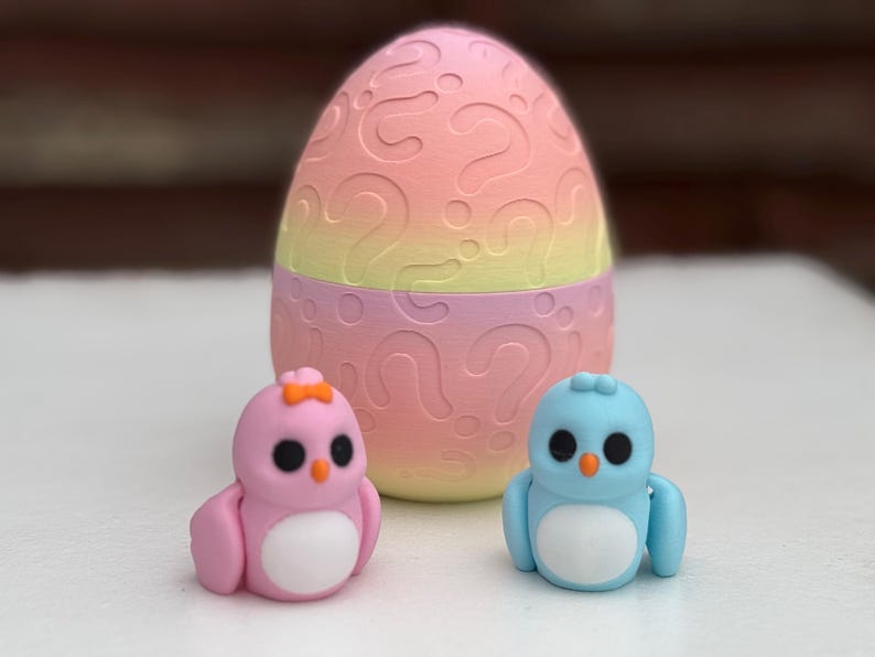 May include: Two small plastic toy birds, one pink and one blue, with white bellies and orange beaks. The birds are sitting in front of a larger pink and yellow egg-shaped toy with question marks on it.