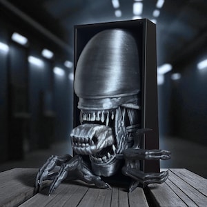 May include: A metallic gray Alien bust sculpture, framed in a black box. The Xenomorph's elongated head, detailed teeth, and extended arms are visible. The sculpture is placed on a wooden surface, with a blurred background.