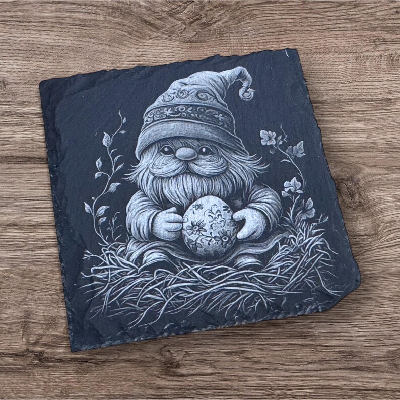Engraved slate coaster featuring a detailed illustration of a bunny resting on a decorative Easter egg, surrounded by grass and leaves. The coaster has a natural, rough-edged finish and is set on a wooden background