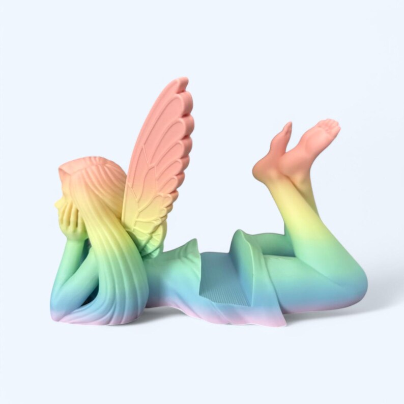 May include: A rainbow-colored fairy figurine with wings lying on its side. The fairy has long hair and is wearing a dress.