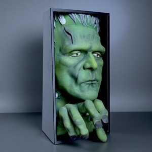May include: A green Frankenstein monster bust emerging from a black framed box. The monster has a green complexion, black hair, and a scar on its forehead. The monster's hands are visible, with dark gray fingernails.