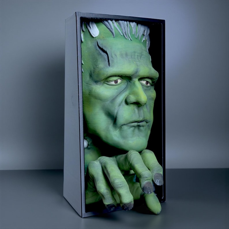 May include: A green Frankenstein monster bust emerging from a black framed box. The monster has a green complexion, black hair, and a scar on its forehead. The monster's hands are visible, with dark gray fingernails.