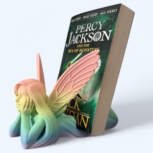 May include: A rainbow-colored fairy bookend holding a book titled "Percy Jackson and the Sea of Monsters". The bookend is made of plastic and has a whimsical design.