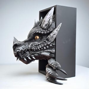 May include: A detailed sculpture of a dragon's head and claws emerging from a black rectangular box. The dragon is primarily gray with silver highlights, a textured surface, and a golden eye. The claws are also gray.