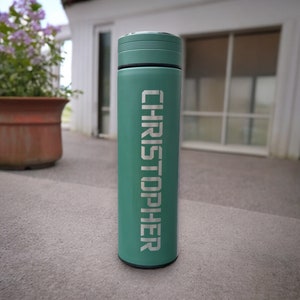 Green vacuum bottle with your name. The bottle is placed on an outdoor patio, with a blurred background featuring a potted plant and a building with windows. The overall setting is casual and outdoor.