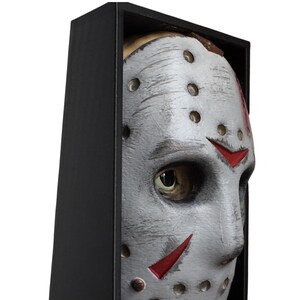May include: A close-up of a white hockey mask with red markings and dark eye holes, set in a black frame. The mask has a weathered appearance with visible scratches and rivets, evoking a horror theme.