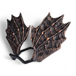 May include: A pair of decorative dragon wings made of a dark brown material with a textured surface. The wings are attached to a black metal stand.