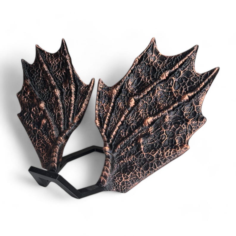 May include: A pair of decorative dragon wings made of a dark brown material with a textured surface. The wings are attached to a black metal stand.