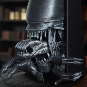 May include: A detailed, metallic gray sculpture of an alien creature emerging from a black rectangular object. The sculpture features intricate details of the alien's head, teeth, and claws, creating a striking and menacing appearance. The overall theme is science fiction and horror.
