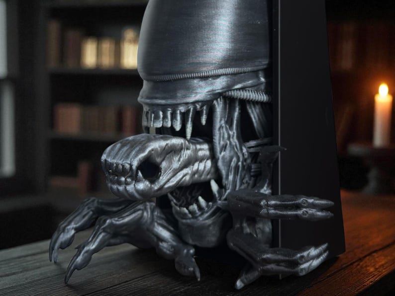May include: A detailed, metallic gray sculpture of an alien creature emerging from a black rectangular object. The sculpture features intricate details of the alien's head, teeth, and claws, creating a striking and menacing appearance. The overall theme is science fiction and horror.