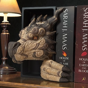 May include: A detailed dragon head bookend in shades of brown, tan, and gold. The dragon's head and claw extend from a black rectangular base, with intricate detailing. The bookend is positioned between two books with the author's name "Sarah J. Maas" visible.