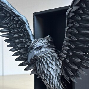 May include: A detailed, gray sculpture of an eagle with outstretched wings. The eagle is positioned within a black, rectangular frame, showcasing intricate feather detailing and a sharp beak. The sculpture is a decorative item.