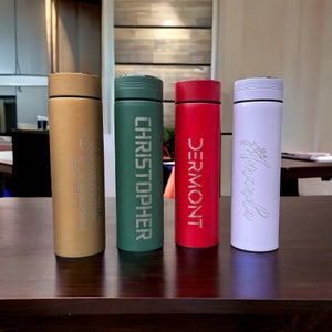 Four colorful thermos bottles with personalized names stand on a table. From left to right: Summer (gold), CHRISTOPHER (green), DERMONT (red), and Amanda (white). The background is an indoor setting with blurred furnishings.
