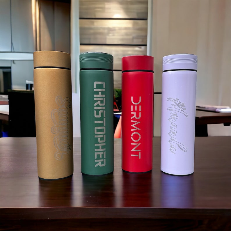 Four colorful thermos bottles with personalized names stand on a table. From left to right: Summer (gold), CHRISTOPHER (green), DERMONT (red), and Amanda (white). The background is an indoor setting with blurred furnishings.