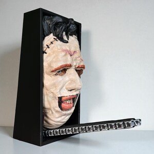 May include: A horror-themed wall decor piece featuring a mask of a character from a horror movie. The mask is set within a black rectangular frame, with a chain-like element extending from the bottom. The mask has a pale complexion, red accents, and black hair.
