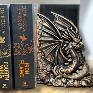 May include: A pair of fantasy novels by Rebecca Yarros titled "Iron Flame" and "Fourth Wing" are propped up by a bronze dragon bookend.