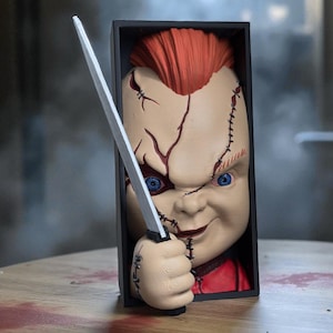 Puede incluir: Figura de mu&ntilde;eco Chucky con pelo rojo y ojos azules, sosteniendo un cuchillo. El mu&ntilde;eco est&aacute; en una caja negra, con puntadas visibles en la cara. El mu&ntilde;eco lleva una camisa roja.