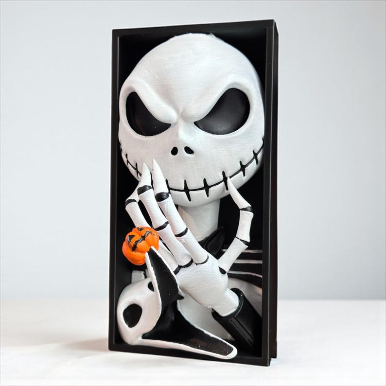 May include: A white Jack Skellington figure with black eyes, a stitched mouth, and a small orange pumpkin. The figure is set in a black rectangular frame, perfect for Halloween decor.