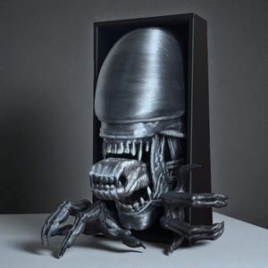 May include: A metallic gray sculpture of an Alien Xenomorph emerging from a black rectangular frame. The sculpture features the creature's elongated head, multiple rows of teeth, and extended claws. The artwork is displayed on a light-colored surface.