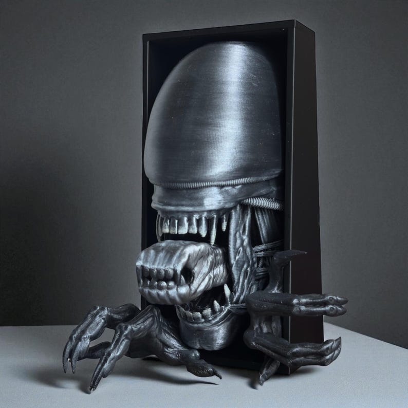 May include: A metallic gray sculpture of an Alien Xenomorph emerging from a black rectangular frame. The sculpture features the creature's elongated head, multiple rows of teeth, and extended claws. The artwork is displayed on a light-colored surface.