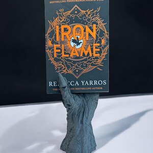 May include: A book titled "Iron Flame" with orange lettering and a black background is held by a gray, textured hand-shaped bookend. The book's cover also features a stylized illustration. The author's name is Rebecca Yarros.