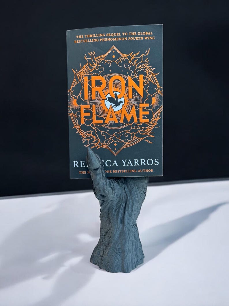 May include: A book titled "Iron Flame" with orange lettering and a black background is held by a gray, textured hand-shaped bookend. The book's cover also features a stylized illustration. The author's name is Rebecca Yarros.